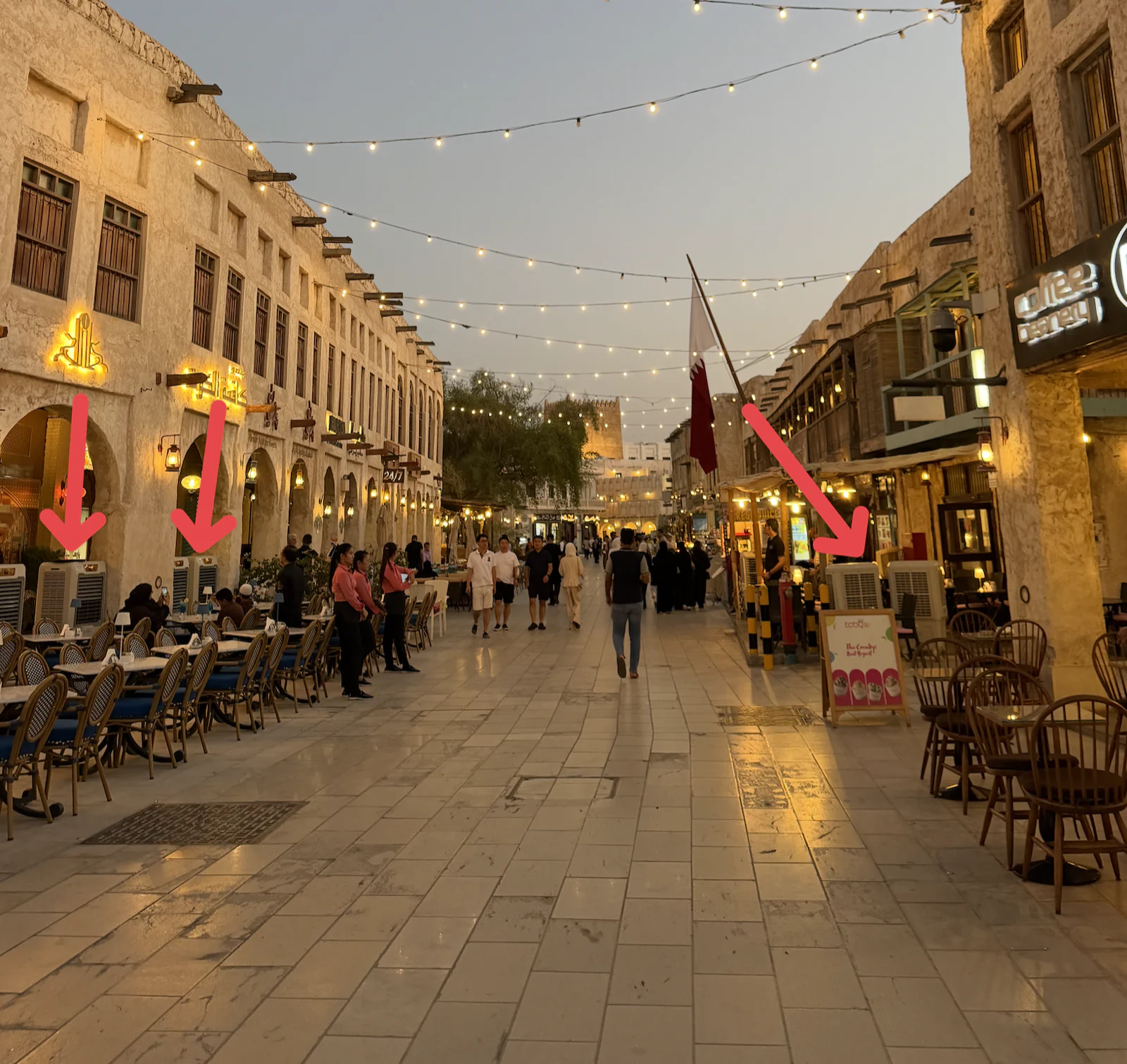 Une allée du souq Waqif à la tombée du jour, guirlandes lumineuses au-dessus des passants et terrasses dressées de part et d'autre. Des flèches rouges désignent trois climatiseurs d'extérieur posés au pied des façades.