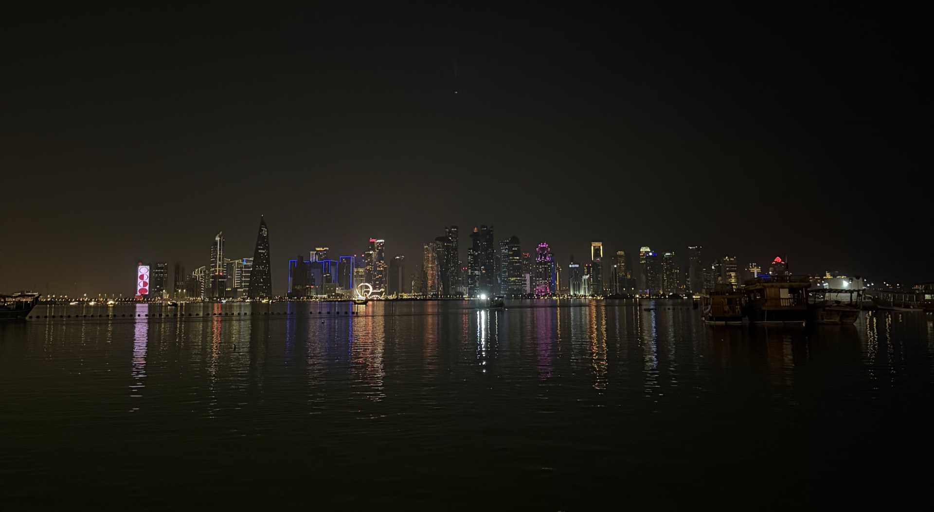 La skyline de West Bay vue depuis la Corniche de Doha, de nuit, avec les boutres amarrés et les reflets des tours sur l'eau.
