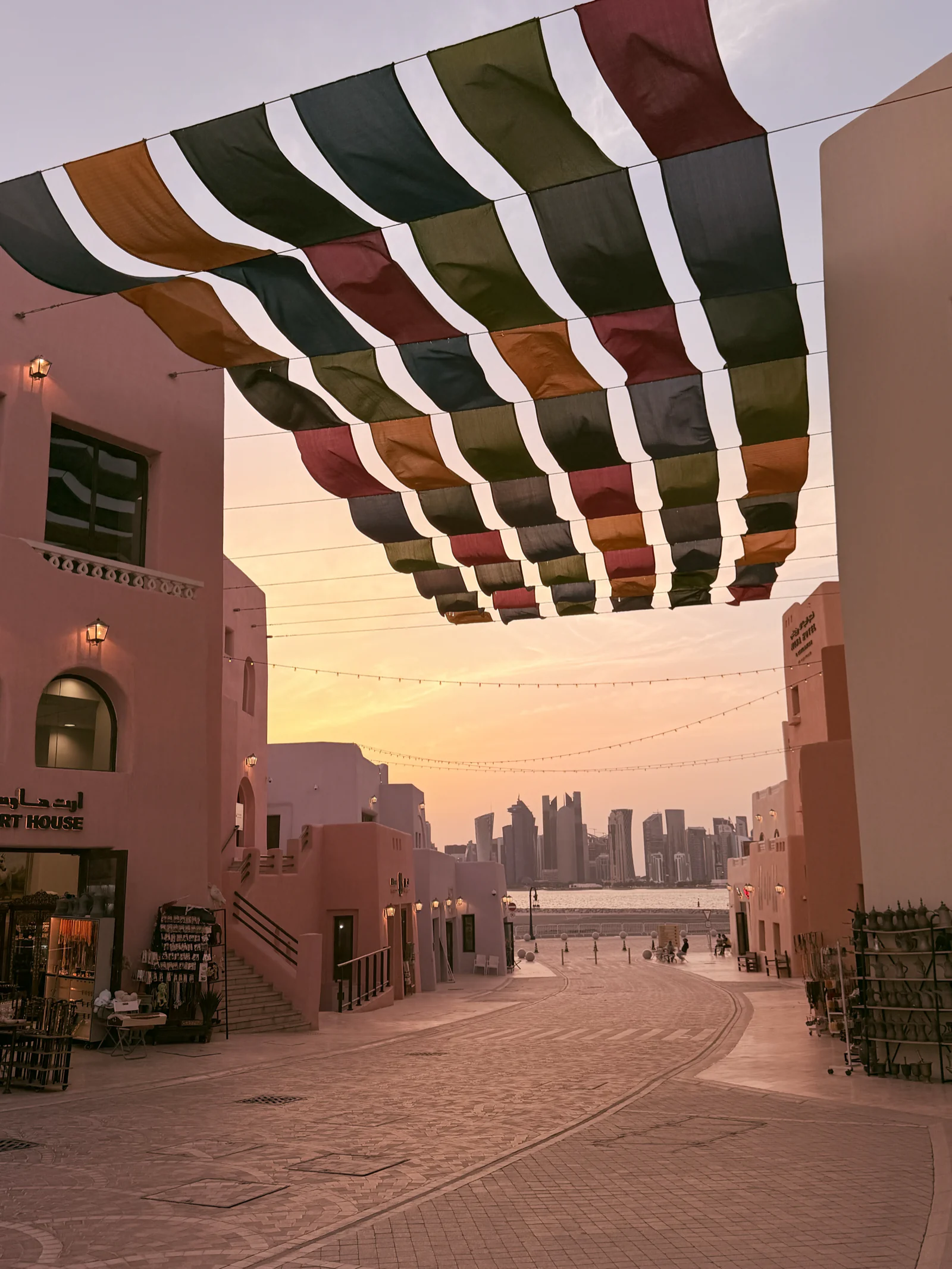 Rue piétonne du vieux port de Doha au coucher du soleil, sous des voiles colorées tendues entre les façades, la skyline de West Bay au fond