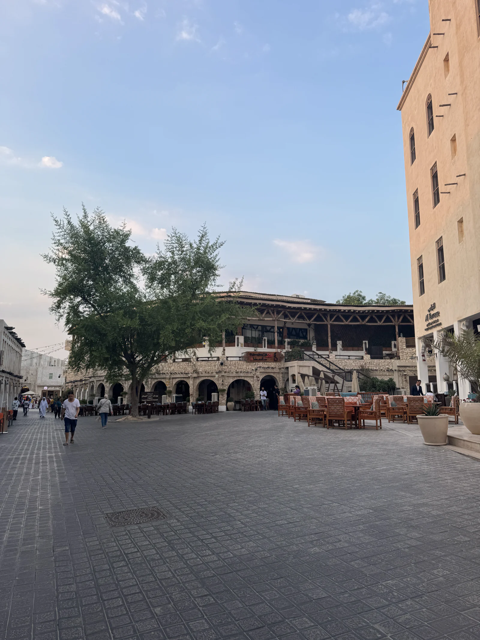 La grande place du souq Waqif en fin de journée, bâtiments beiges et terrasses à l'ombre d'un arbre