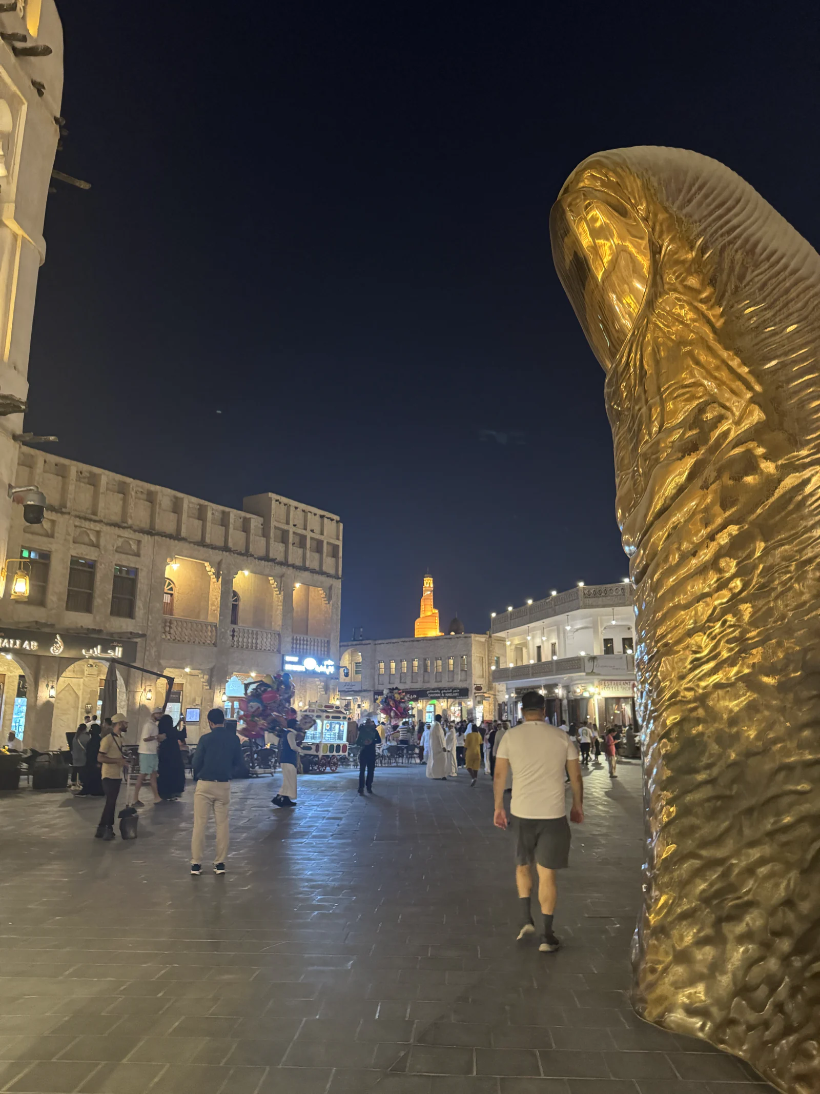 Le Pouce doré de César sur une place du souq Waqif la nuit, la tour du souq éclairée au fond