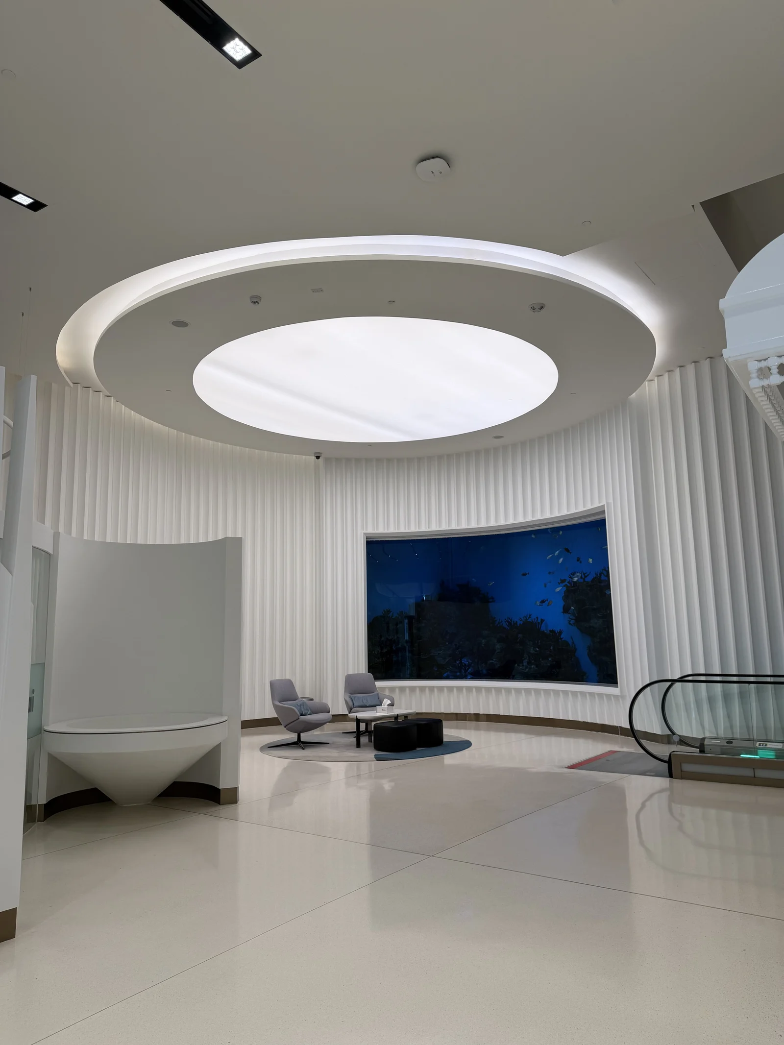 Un salon d'attente circulaire sous un plafond lumineux, écran mural et fauteuils